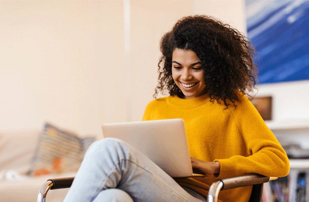 A woman with curly hair and a yellow sweater sits with her laptop, accessing her MySelective online account,